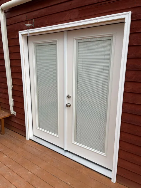 White double doors with decorative glass panels, set in a red exterior wall, on a wooden deck.